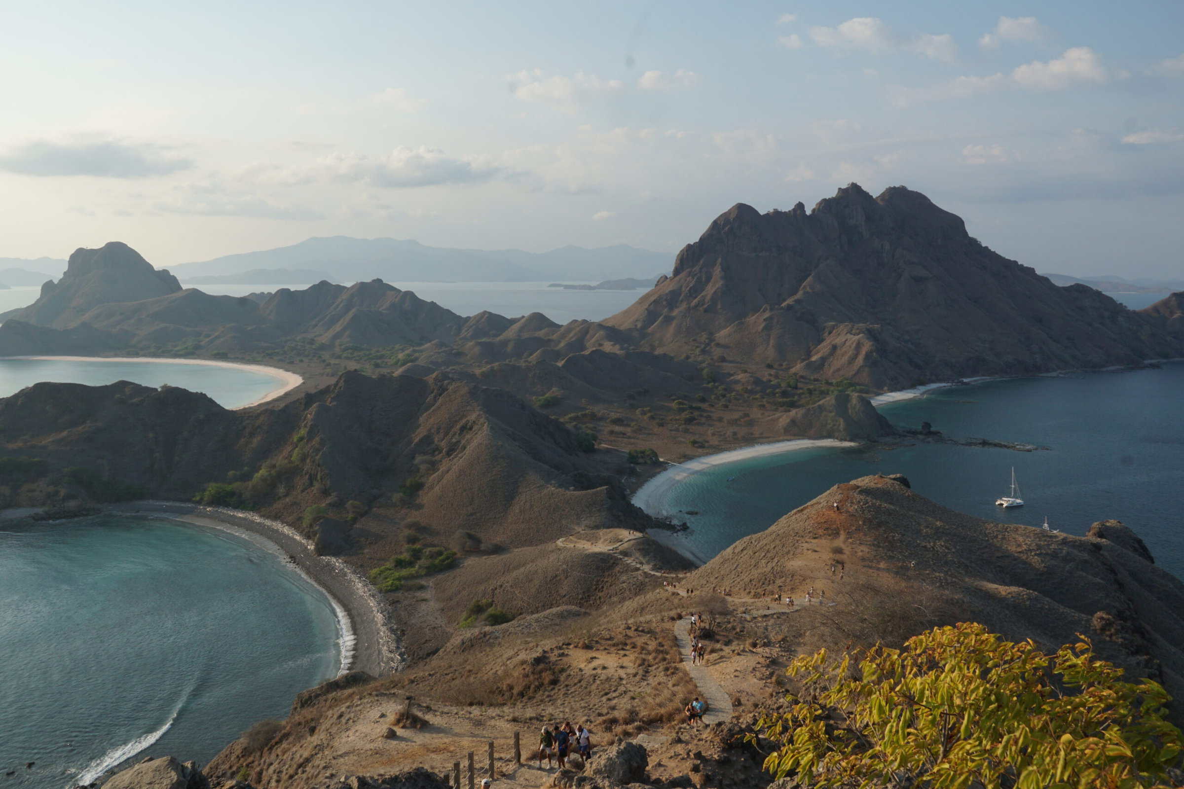 A scenic view of a rugged coastal landscape with curving beaches, turquoise waters, and rocky hills under a clear sky, where a few people roam along a trail in the foreground, pausing to rest and take in the beauty.