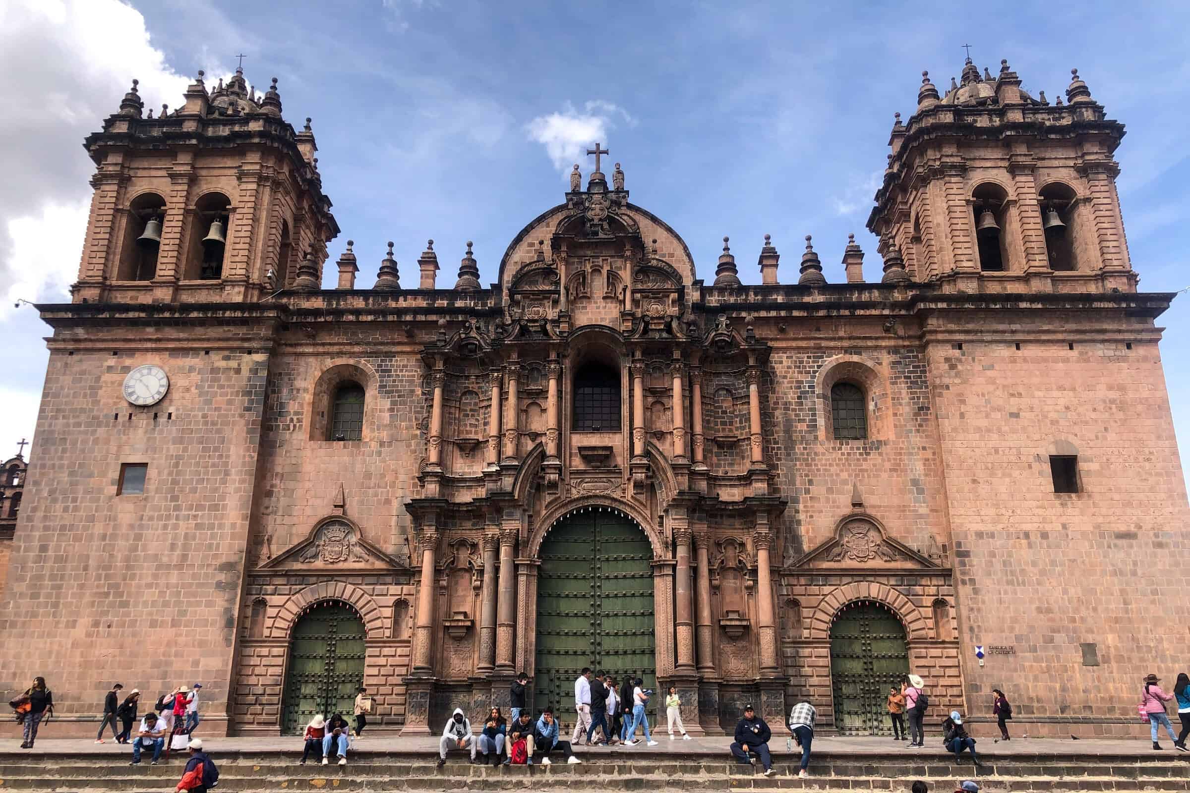 Large stone cathedral with ornate baroque facade, arched doors, and twin towers, situated on a wide stone staircase. People are sitting and walking on the steps under a blue sky.