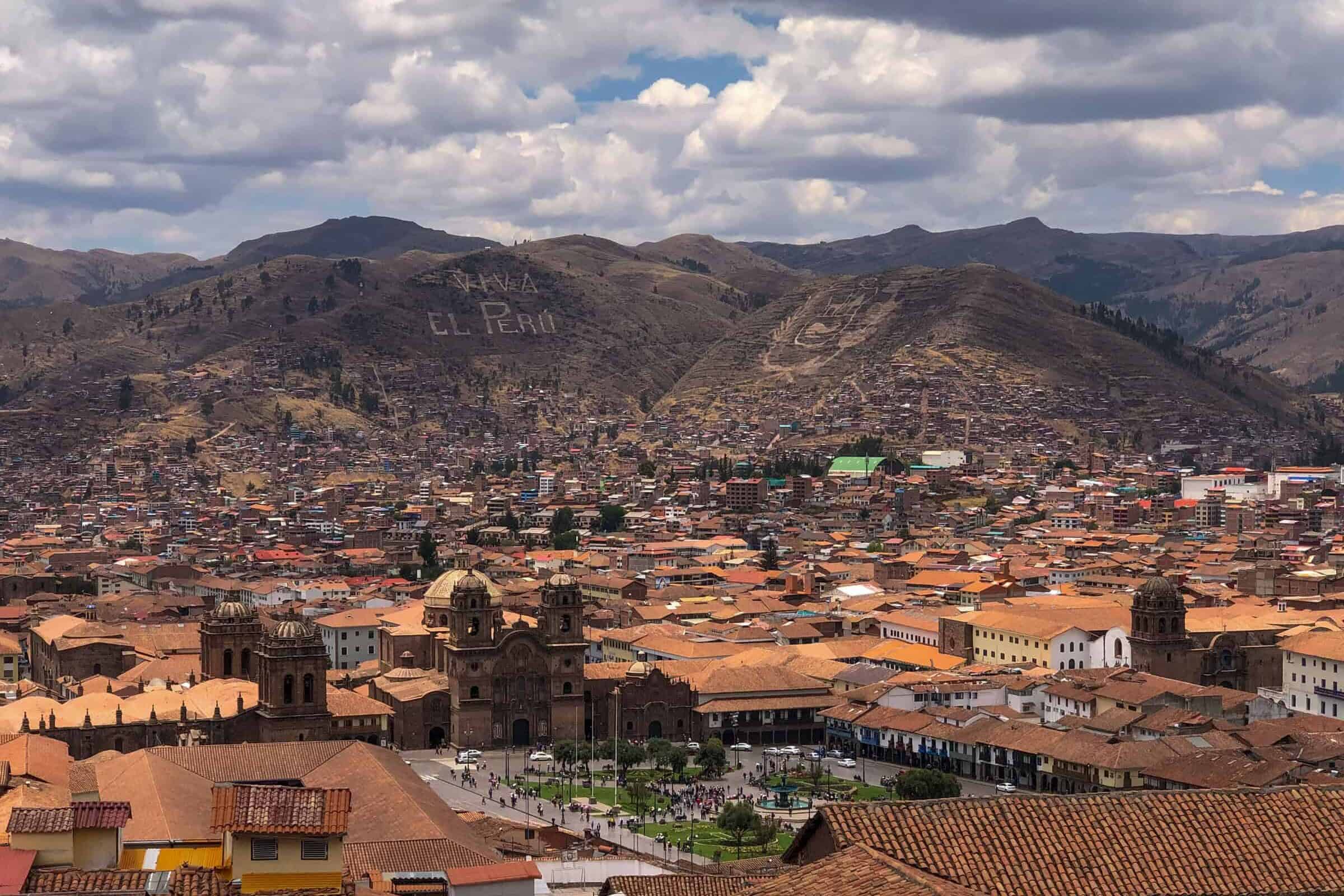 A panoramic view of Cusco, Peru, featuring red-tiled rooftops, colonial buildings, and surrounding mountains with “Viva el Perú” written on a hillside under a partly cloudy sky.