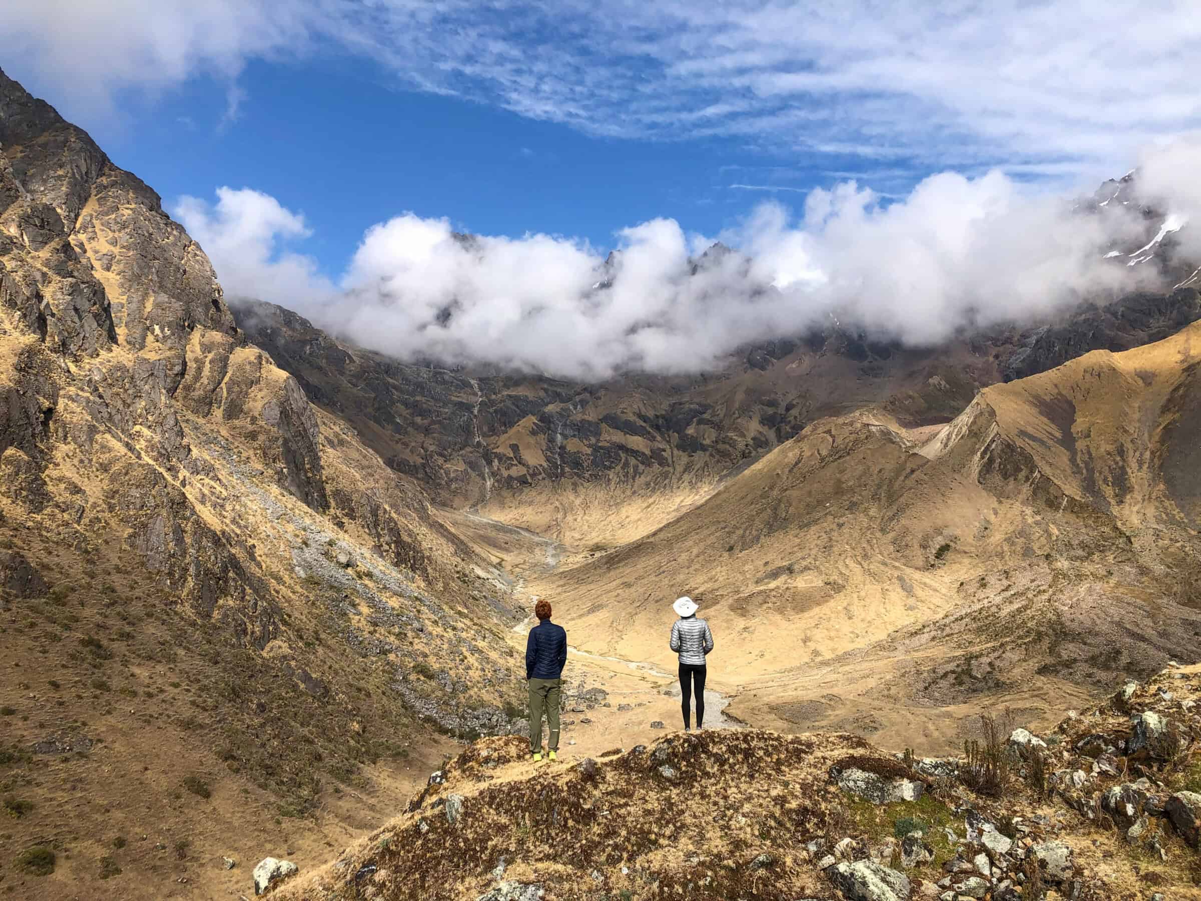 Two people stand on a rocky hilltop overlooking a vast, dry mountain valley with rugged peaks and scattered clouds beneath a blue sky. The landscape is golden brown with sparse vegetation.