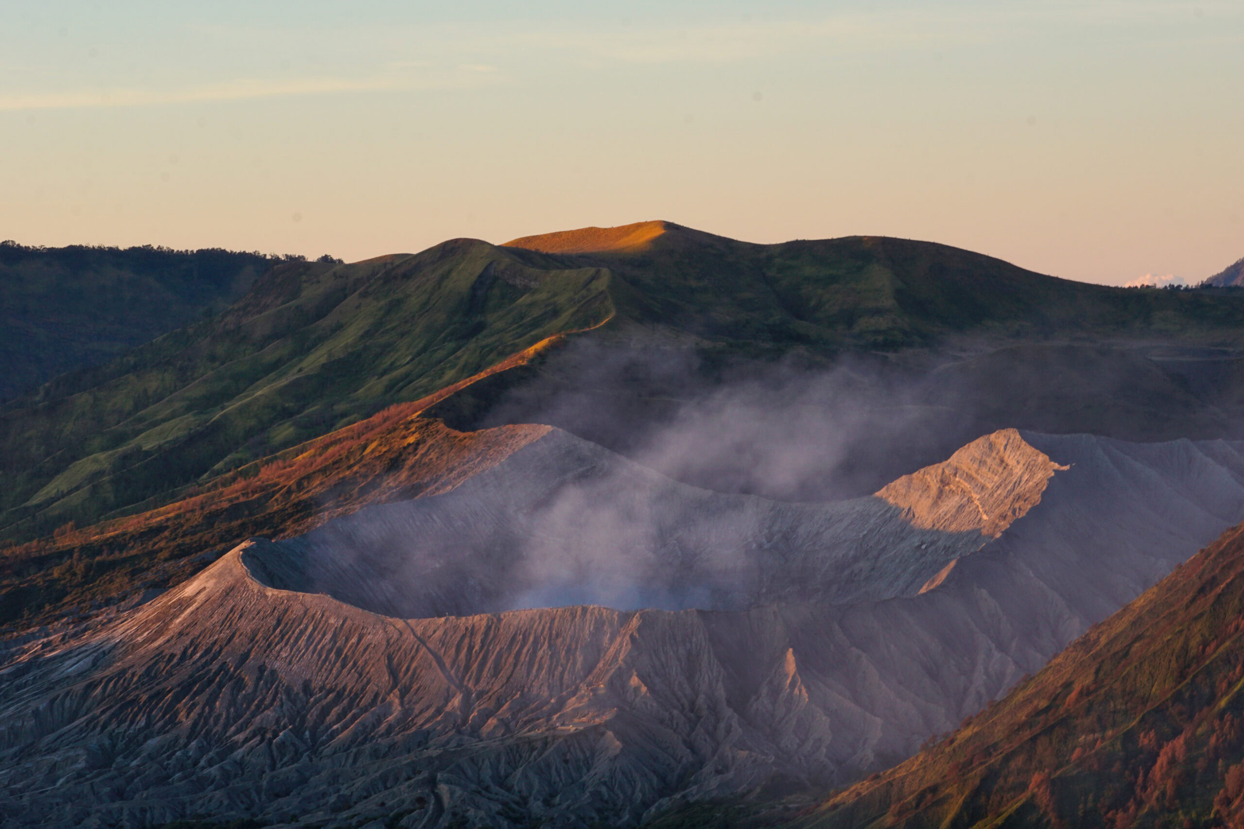 A smoking volcanic crater surrounded by rugged, green hills is bathed in soft sunlight under a clear sky. Wisps of steam rise from the crater, highlighting the textured, grayish surface.