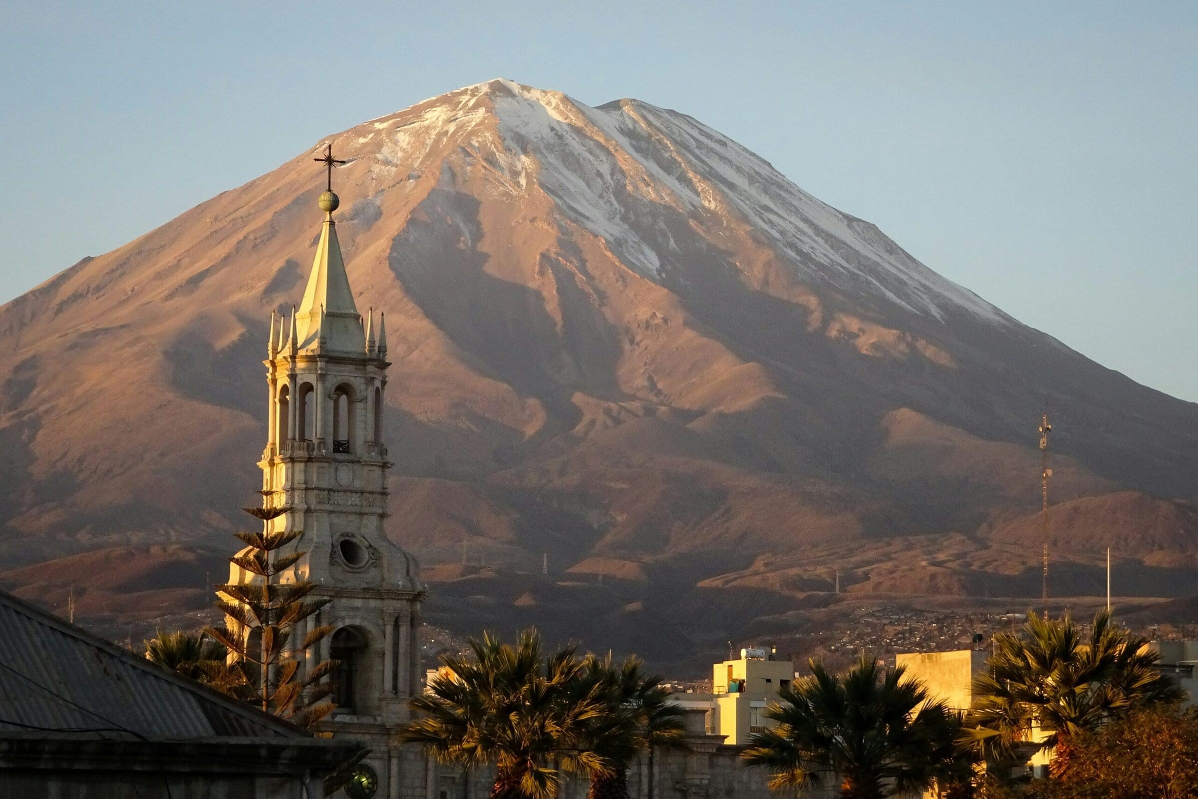 A tall church tower with a cross stands in front of a snow-capped mountain, with palm trees and buildings at the base, all lit by the golden light of sunset.