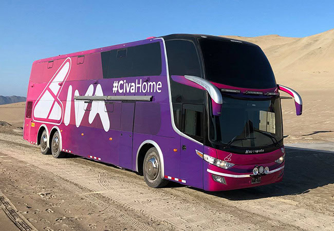 A large purple and pink double-decker bus with the text #CivaHome and CIVA on its side is parked on a sandy road in a desert landscape under a clear blue sky.