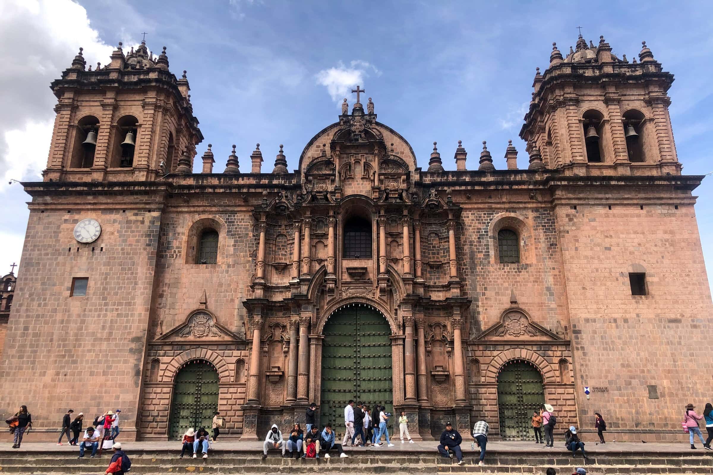 A large stone cathedral with ornate details and twin towers stands behind wide steps, where groups of people are sitting and walking; the sky is partly cloudy.