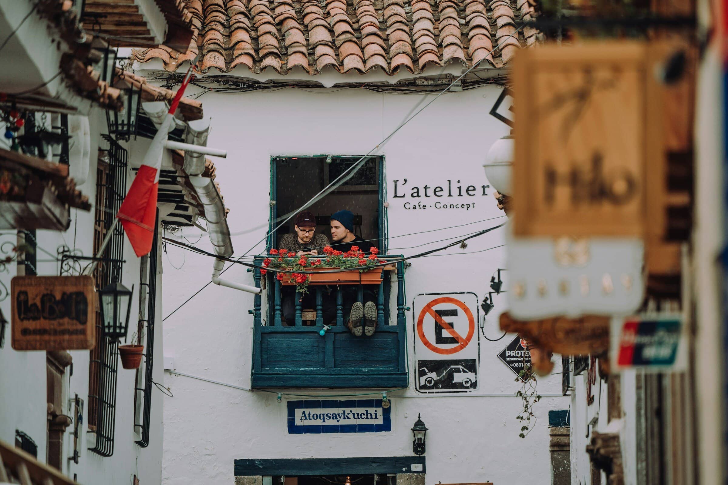 Two people sit on a small blue balcony with red flowers, above a narrow street lined with hanging signs and lanterns. The white building has a sign reading L’atelier Café Concept. The atmosphere is cozy and quaint.