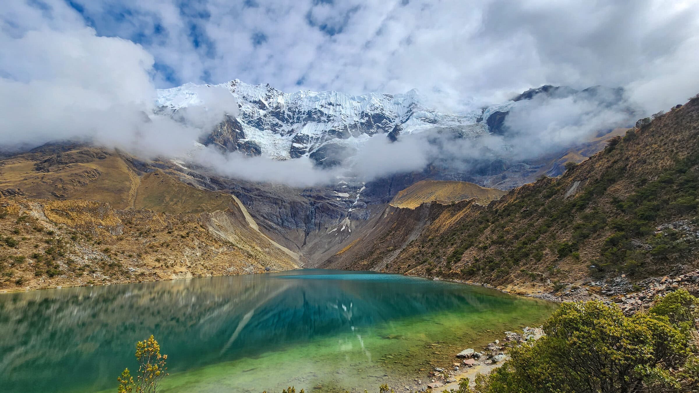 A turquoise lake surrounded by rocky hills and green shrubs, with snow-capped mountains towering in the background and clouds partially covering the peaks under a partly cloudy sky.