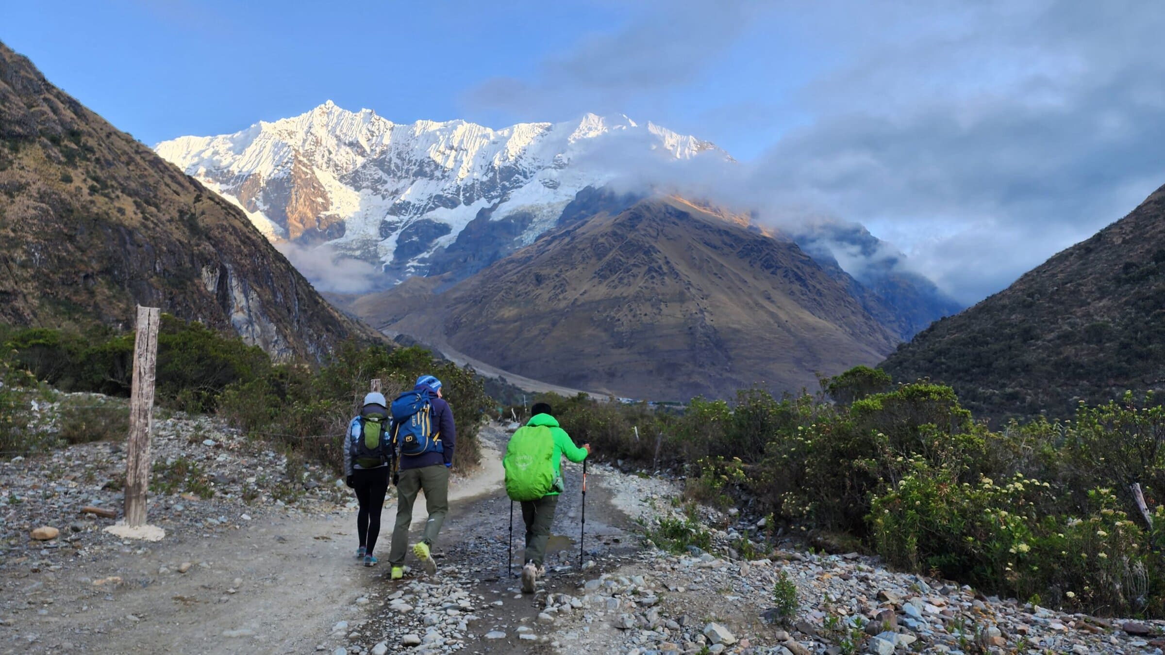 Three hikers with backpacks walk along a rocky mountain trail, surrounded by greenery, with snow-capped peaks and a partly cloudy sky in the background.