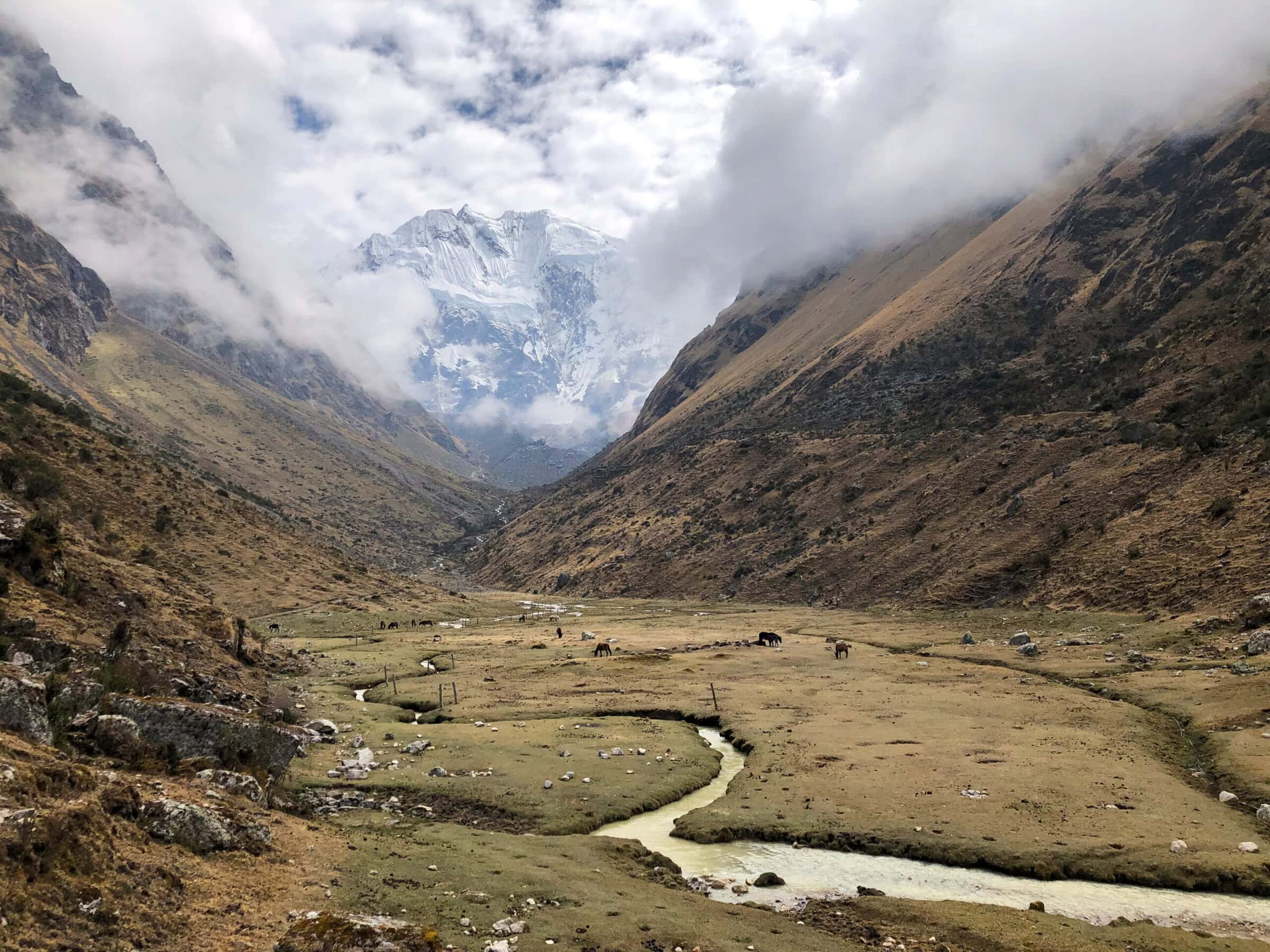 A winding stream flows through a grassy valley with grazing animals, flanked by steep, brown mountains. Snow-capped peaks rise in the background beneath a partly cloudy sky.