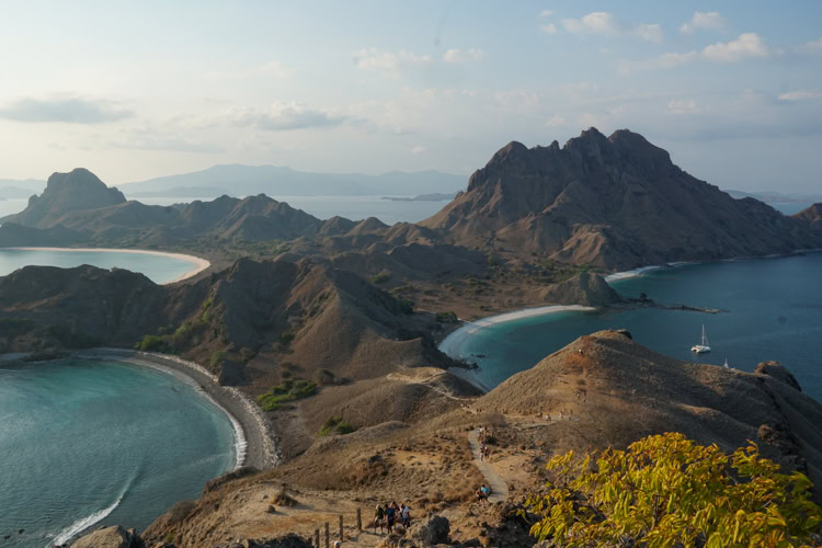 A panoramic view of Padar Island in Indonesia, showing rugged brown hills, curved sandy beaches, and turquoise blue waters under a partly cloudy sky. A few people are visible on a trail near the foreground.