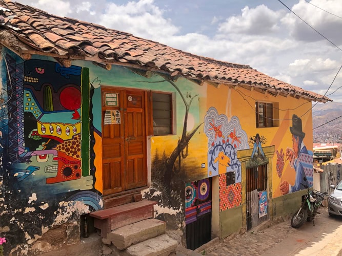 A colorful mural covers the side of a rustic building with a tiled roof, featuring vibrant abstract and figurative art. A wooden door and windows are visible, with a motorcycle parked on the cobblestone street outside.